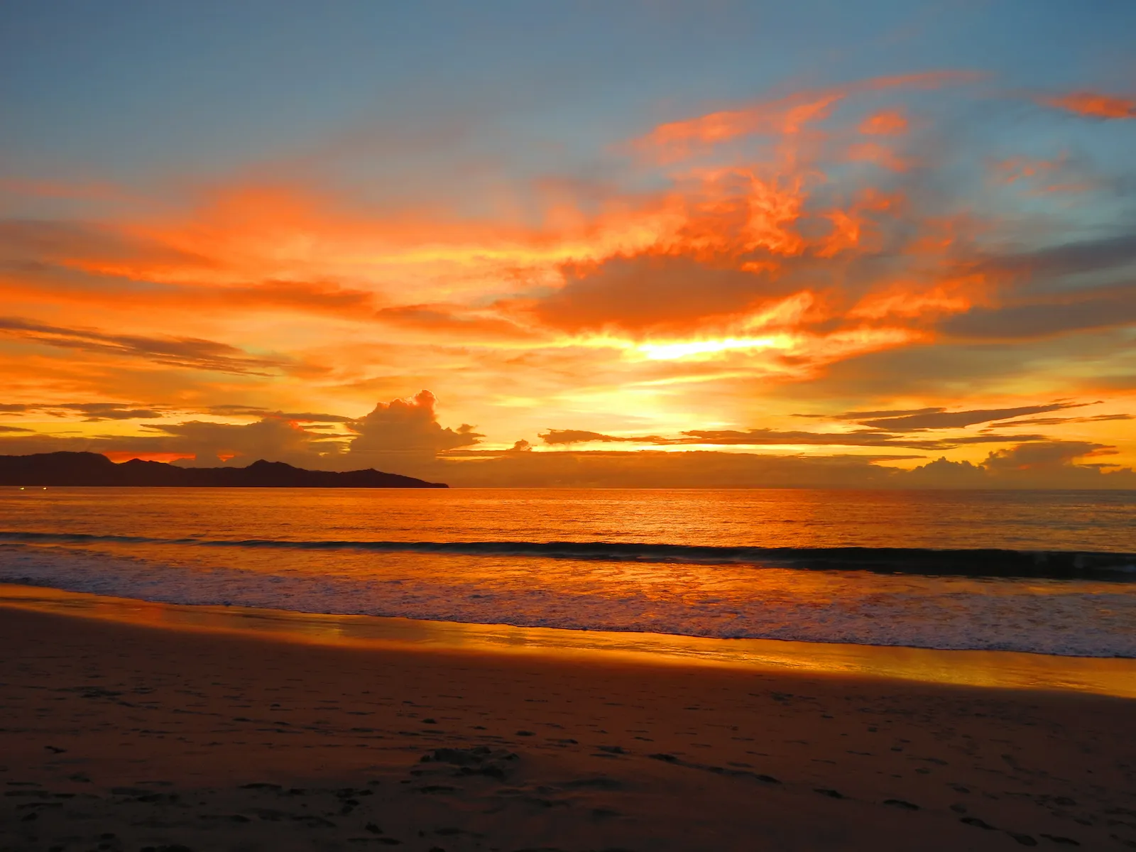 Vibrant sunset over a Costa Rican beach with orange clouds, gentle waves and a rocky silhouette offshore.